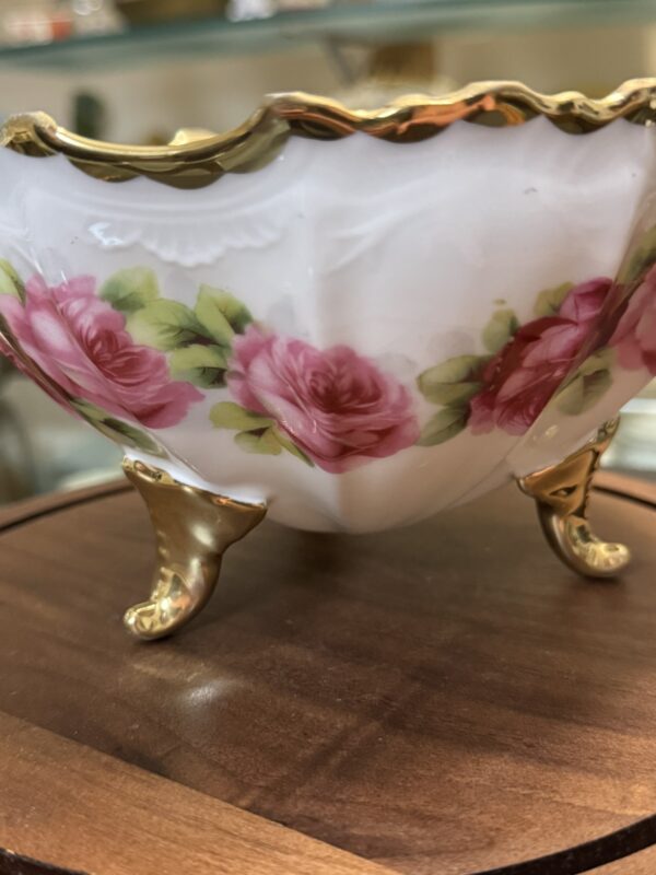 A close-up of a decorative porcelain bowl with scalloped gold trim, ornate gold legs, and a floral design featuring pink roses with green leaves. The bowl is displayed on a wooden surface, with blurred shelves in the background.