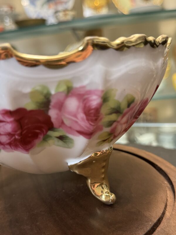 A porcelain bowl with gold trim and ornate gold legs sits on a wooden surface. The bowl is decorated with pink and red roses with green leaves. The background shows glass shelves with blurred decorative items on display.