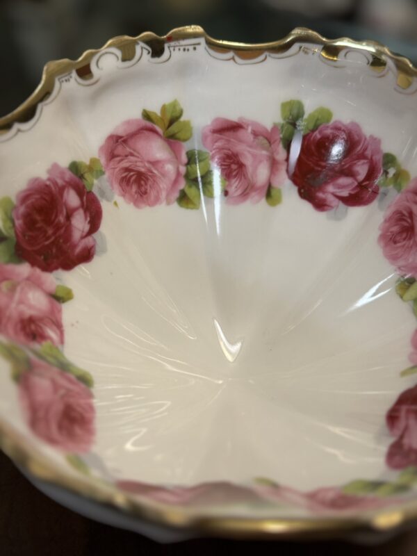 A close-up of a scalloped, white porcelain bowl with a gold-trimmed rim. The inner edge is decorated with a garland of pink and red roses with green leaves. The glossy glaze reflects light, highlighting the elegant floral design.