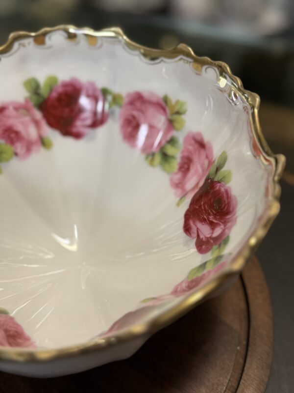 A close-up of a white, scalloped-edge porcelain bowl with a gold rim and pink and red rose patterns along the inner sides. The bowl rests on a dark wooden surface, and the background is softly blurred.