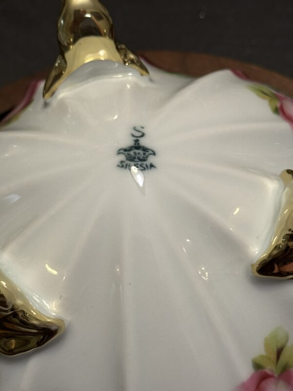Close-up of the underside of a porcelain dish with a white, fluted base, gold accents, and pink floral designs. At the center is a dark blue maker’s mark with a crown, an “S,” and the word “SILESIA” printed below.