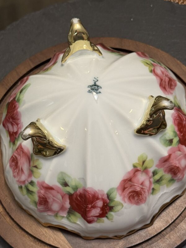 A porcelain dish turned upside down displays gold-accented feet, hand-painted pink and red roses with green leaves, and the “Saji China Japan” maker’s mark in blue. The dish rests on a round wooden surface.