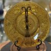 A decorative amber glass plate with intricate floral and geometric patterns is displayed upright on a black metal stand. The background shows other glassware slightly out of focus on glass shelves. The plate sits on a round wooden base.