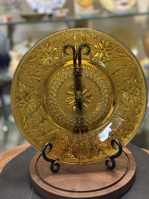 A decorative amber glass plate with intricate floral and geometric patterns is displayed upright on a black metal stand. The background shows other glassware slightly out of focus on glass shelves. The plate sits on a round wooden base.