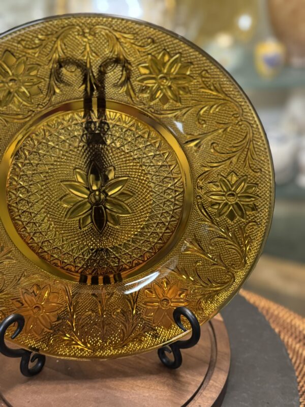 A decorative amber glass plate with intricate floral and geometric patterns is displayed upright on a black metal stand. The plate features detailed embossed flower designs and ornate swirls, set against a blurred background of various light colors.