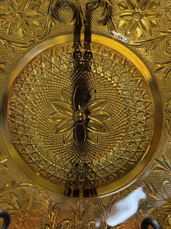 Close-up of an ornate amber glass plate with a detailed floral and geometric pattern. The center features a large flower with six petals, surrounded by textured, radiating designs and smaller flowers along the border. Light reflects off the raised elements.