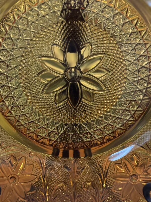 A close-up photo of ornate amber glass featuring a raised floral pattern in the center. The design includes a large flower surrounded by textured, geometric details and additional floral motifs around the edges, all highlighted by the warm, golden color of the glass.