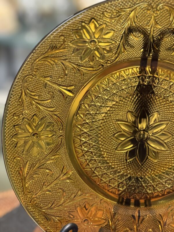 A close-up of an ornate amber glass plate with intricate floral and geometric patterns. The plate features embossed flowers and scroll designs around the rim, with a textured, radiating central motif. Light reflects through the translucent yellow glass.
