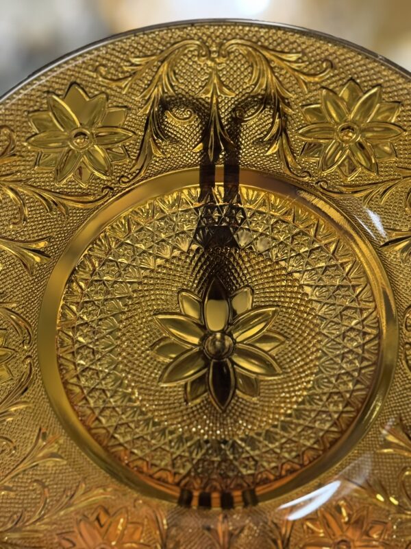 A close-up of an ornate amber glass plate features intricate geometric and floral patterns, including prominent embossed flowers and textured designs that catch the light, creating a warm, glowing effect. The plate’s translucent surface enhances its decorative details.