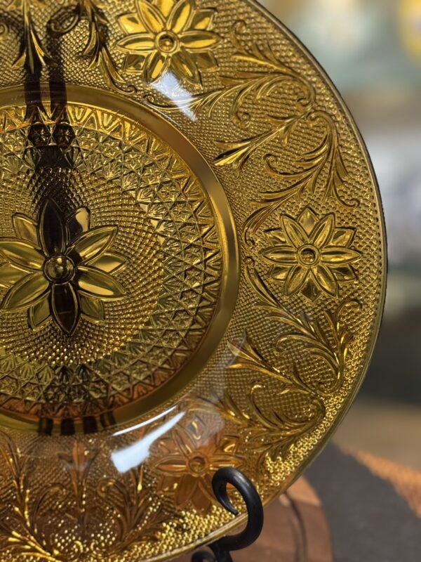 A close-up of an ornate amber glass plate with intricate floral and geometric embossed patterns. The plate is held upright on a black stand, with bright lighting highlighting its translucent amber color and detailed design. The background is softly blurred.