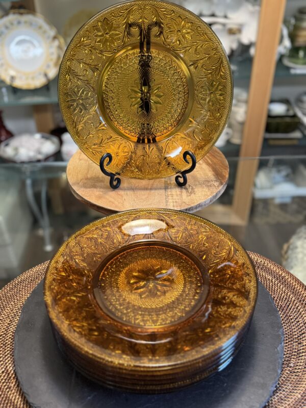 A stack of amber-colored vintage glass plates with intricate floral and geometric patterns sits on a table; one plate is displayed upright on a stand behind the stack, set in a glass cabinet with more dishware in the blurred background.