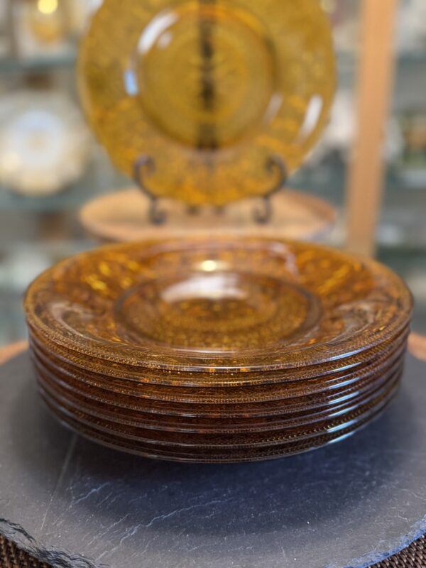 A stack of seven amber-colored glass plates with intricate patterns sits on a dark round surface. In the background, a similar plate is displayed upright on a stand, with shelves of glassware and tableware softly blurred behind it.