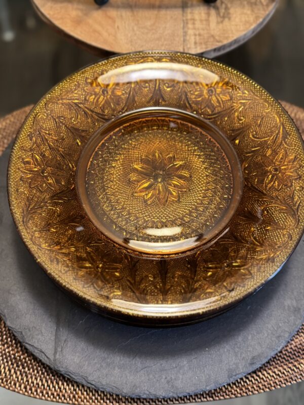 A decorative amber glass plate with an intricate floral and geometric pattern is displayed on a round black slate platter atop a brown woven placemat. The plate’s detailed design reflects light, highlighting its vintage and ornate appearance.