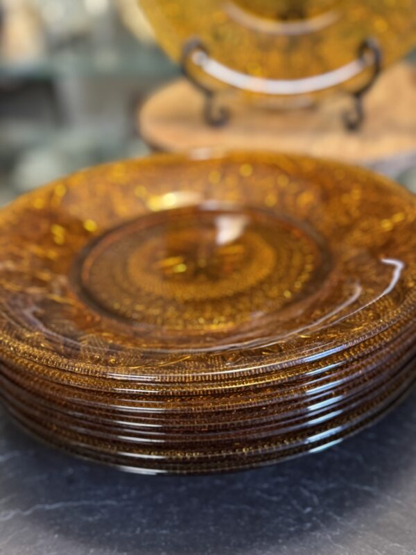 A stack of five translucent amber glass plates with intricate textured patterns sits on a dark countertop. Another similar plate is displayed upright on a stand in the softly blurred background. The glass has a warm, golden-brown hue.