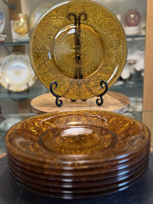 A set of amber glass plates with intricate floral and geometric patterns. One plate is displayed upright on a black stand, while six matching plates are stacked in front. Decorative china is visible in a glass cabinet background.