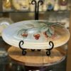 An oval ceramic plate with a hand-painted design of red cherries and green leaves is displayed on a black metal stand. The plate sits on a round wooden surface, with shelves of decorative dishes and ceramics blurred in the background.