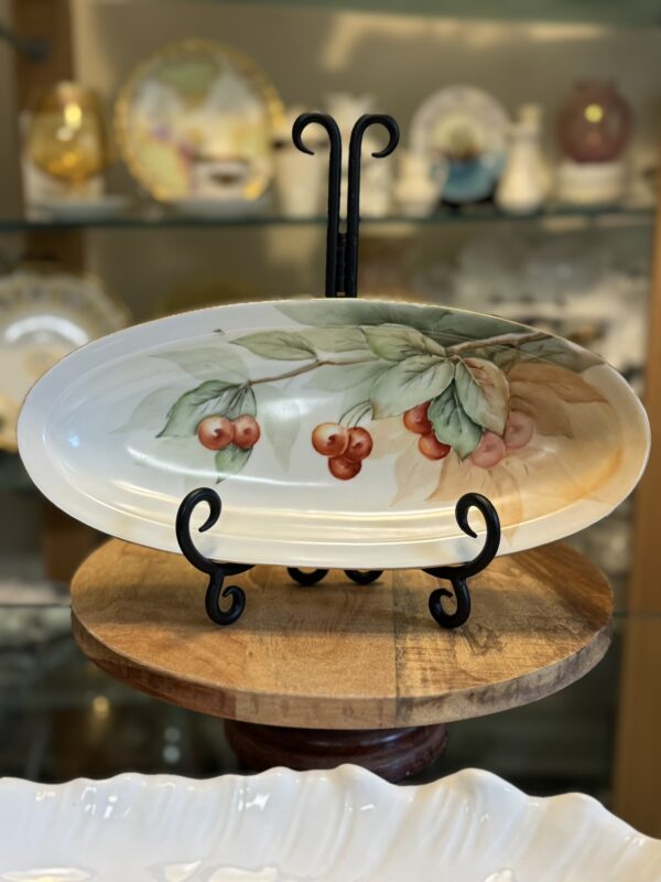 An oval ceramic plate with a hand-painted design of red cherries and green leaves is displayed on a black metal stand. The plate sits on a round wooden surface, with shelves of decorative dishes and ceramics blurred in the background.