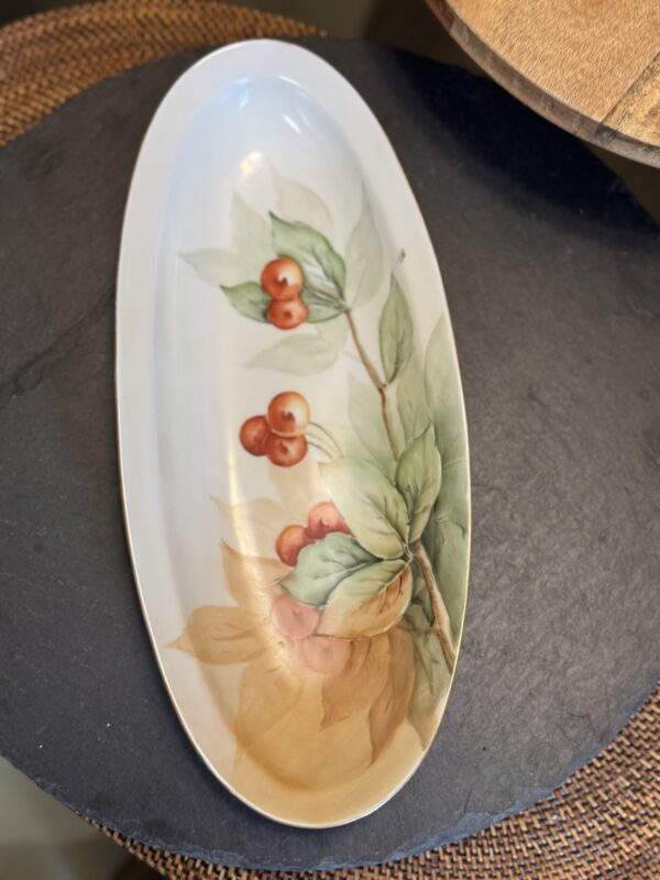 An oval ceramic platter with a hand-painted design of red cherries and green leaves on one side. The plate has a glossy finish and rests on a dark, textured surface with a woven mat partially visible underneath.