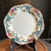 An ornate porcelain plate with an octagonal shape is displayed on a black stand. The plate features a colorful floral border with blue, pink, yellow, and green patterns, set against a white center. The background is softly blurred and dark-toned.