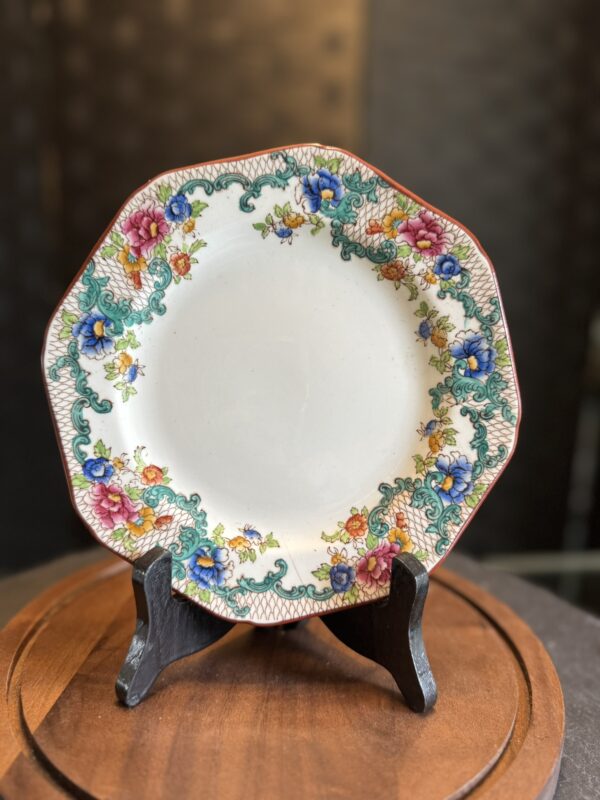 An ornate porcelain plate with an octagonal shape is displayed on a black stand. The plate features a colorful floral border with blue, pink, yellow, and green patterns, set against a white center. The background is softly blurred and dark-toned.