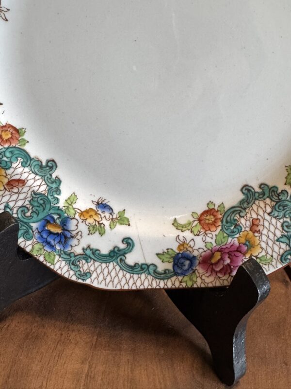 A decorative ceramic plate with a scalloped edge features a border of colorful floral patterns and green scrollwork over a gold mesh background. The plate rests on a black display stand on a wooden surface. A small chip is visible on the edge.