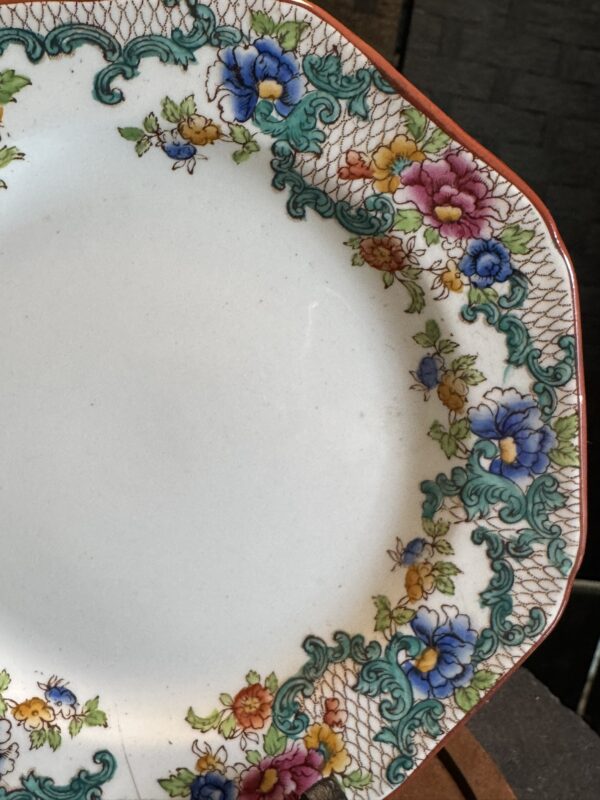 A white porcelain plate with scalloped edges, decorated with a colorful floral and green scrollwork border. The rim features red trim, and clusters of blue, pink, purple, and yellow flowers with green leaves around the edge.