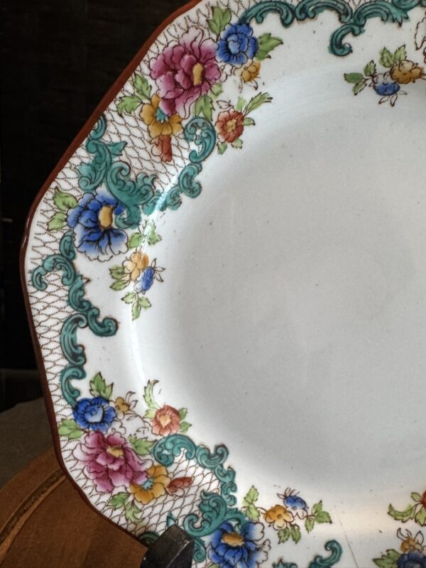 A close-up of a vintage ceramic plate with a floral pattern. The edge features ornate teal scrollwork and clusters of colorful flowers in pink, blue, yellow, and red on a white background, bordered by a brown line. Part of the plate stand is visible.