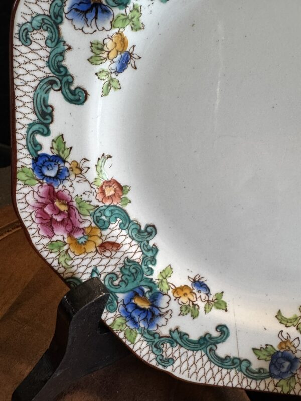 A close-up of a decorative ceramic plate with a floral pattern along the edge. The flowers are pink, yellow, blue, and purple, surrounded by green ornamental swirls and a brown lattice design. The plate is displayed on a dark wooden stand.