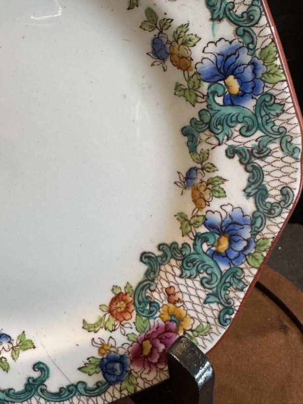 A close-up of a decorative plate’s edge, featuring a floral and ornate teal scroll border with blue, purple, yellow, and red flowers. The background is white, and the plate stands on a dark holder. Delicate net-like patterns accent the rim design.