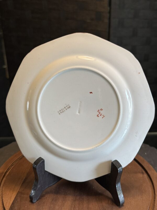 A small, white, octagonal ceramic plate is displayed upright on a dark wooden stand. The plate’s underside is visible, showing a stamp that reads “FALCON CHINA ENGLAND” and some red handwritten marks. The background is softly blurred and dark.