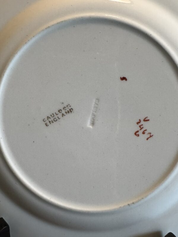 The underside of a white ceramic plate shows three markings: Cauldon England stamped in black, an indented mark at the center, and handwritten red numbers and letters reading V 2467 2. The surface appears clean and slightly glossy.