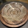 A clear, intricately patterned glass bowl with a diamond-cut design sits on a round wooden tray. The tray has a natural wood grain and two contrasting segments. The background is slightly out of focus, emphasizing the bowl and tray.