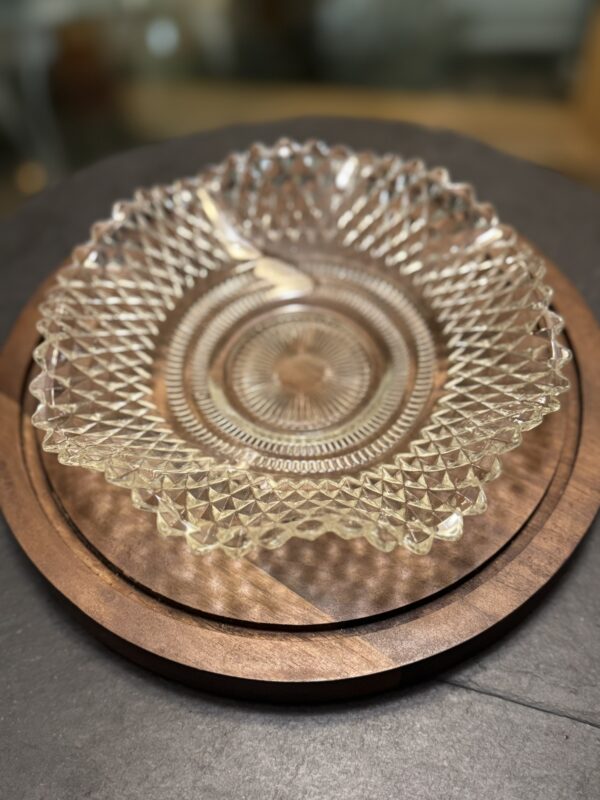 A clear, intricately patterned glass bowl with a diamond-cut design sits on a round wooden tray. The tray has a natural wood grain and two contrasting segments. The background is slightly out of focus, emphasizing the bowl and tray.