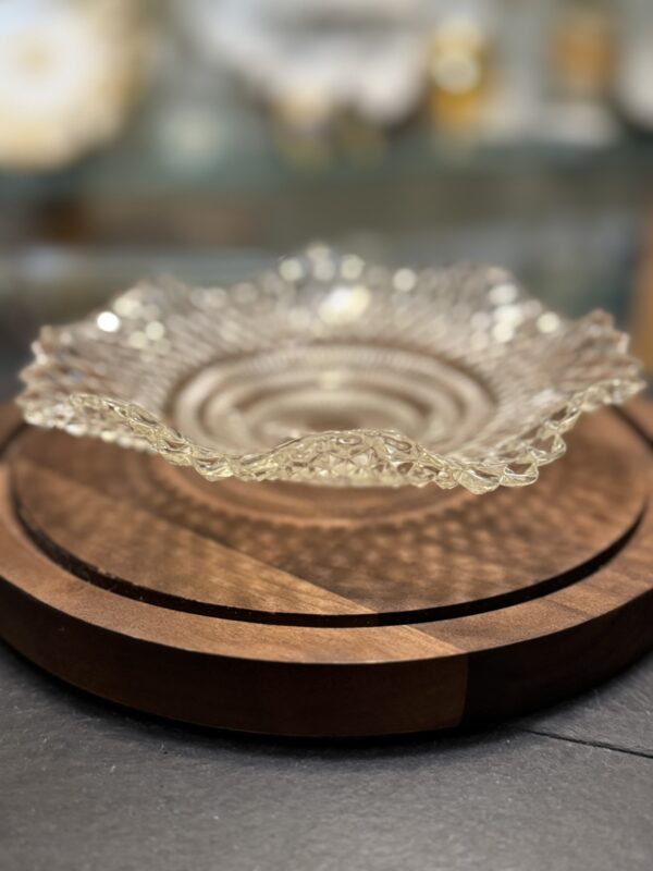 A clear, scalloped glass plate with a textured, intricate pattern sits on a round, two-tone wooden tray. The background is softly blurred, highlighting the plate’s delicate details and the tray’s smooth finish.