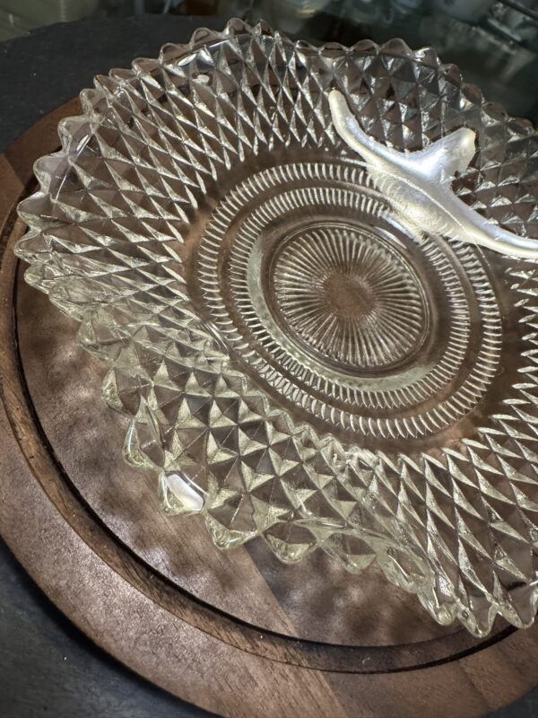 A clear, vintage glass dish with a textured, diamond-patterned rim and a small curved glass handle sits on a round wooden board. The dish catches light, emphasizing its intricate details. The background is dark and slightly out of focus.
