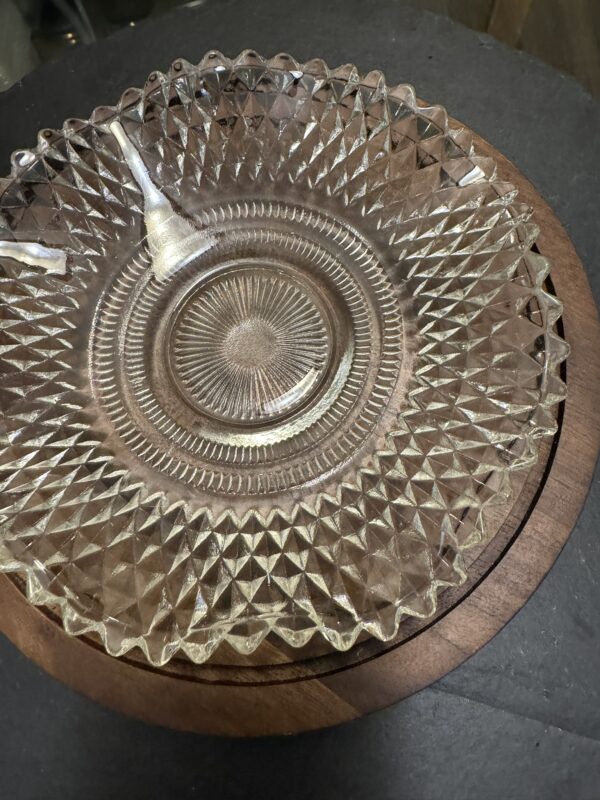 A clear, glass plate with a diamond-patterned edge and sunburst center sits on a round wooden tray. Two small glass serving spoons rest inside the plate, and the setup is placed on a dark surface.