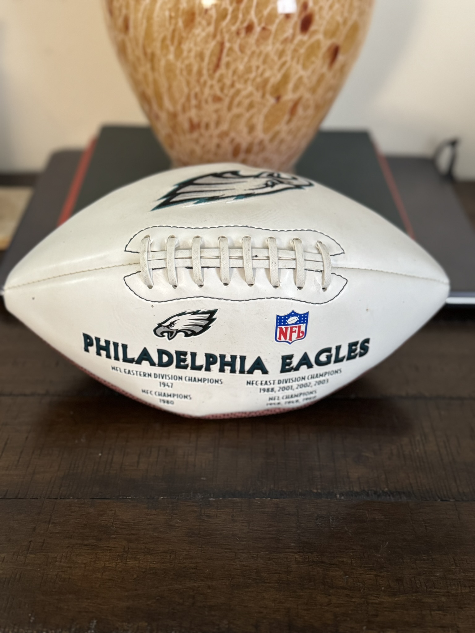 A commemorative white Jeremiah Trotter, Sr. Philadelphia Eagles Football featuring the team and NFL logos sits on a wooden surface, with a blurred orange vase and dark book in the background.