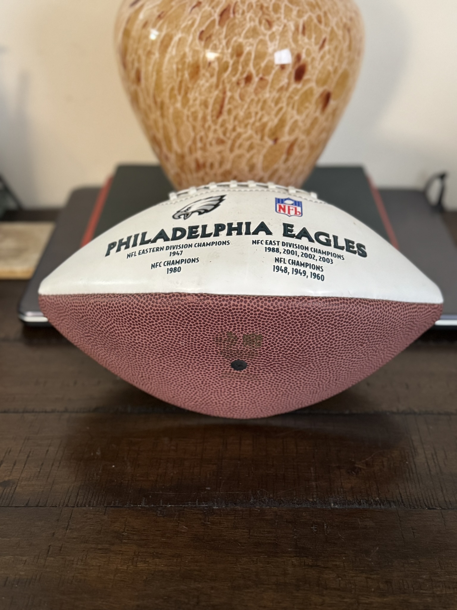 A Jeremiah Trotter, Sr. Philadelphia Eagles Football rests on a wooden table, its white top half showing the team name, logo, NFL logo, and championship years. Behind it is a brown lamp on a closed laptop—an ideal display for fans.