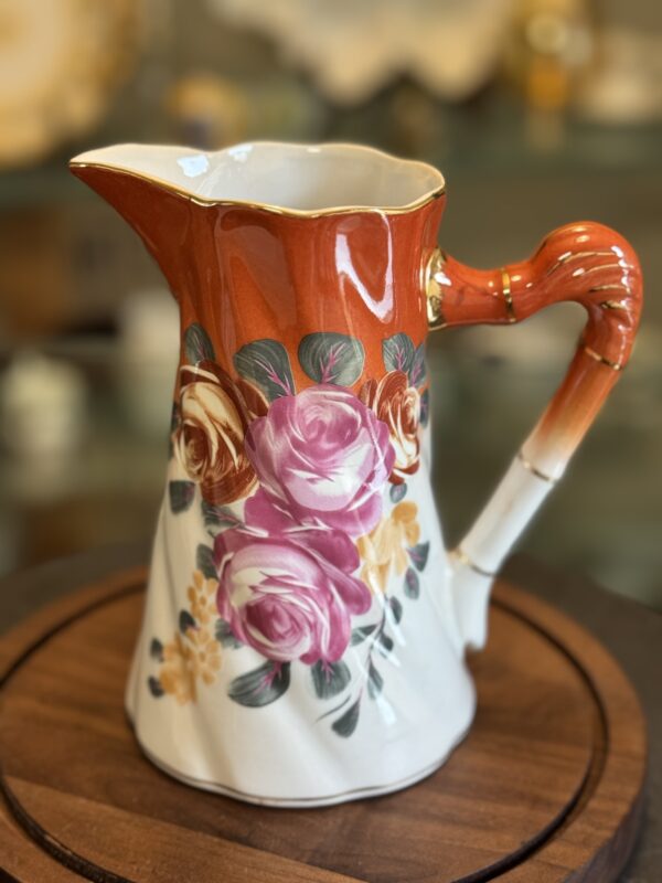 The Vintage Floral Pitcher features a curved handle, glossy orange top, and white base with painted pink, yellow, and white roses with green leaves. This ceramic pitcher is displayed on a round wooden surface against a blurred background.
