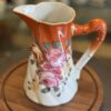The Vintage Floral Pitcher features a glossy orange top and handle, decorated with pink and yellow roses. This ceramic piece is displayed on a round wooden tray, with various ceramics softly blurred in the background.