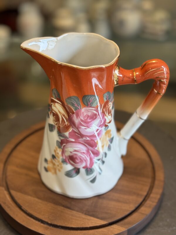 The Vintage Floral Pitcher features a glossy orange top and handle, decorated with pink and yellow roses. This ceramic piece is displayed on a round wooden tray, with various ceramics softly blurred in the background.