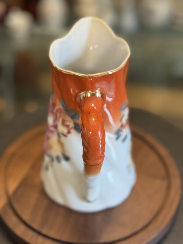 The Vintage Floral Pitcher features an ornate curved handle and glossy finish, with an orange top trimmed in gold, a white base adorned with painted flowers, and rests on a round wooden surface against a blurred background.