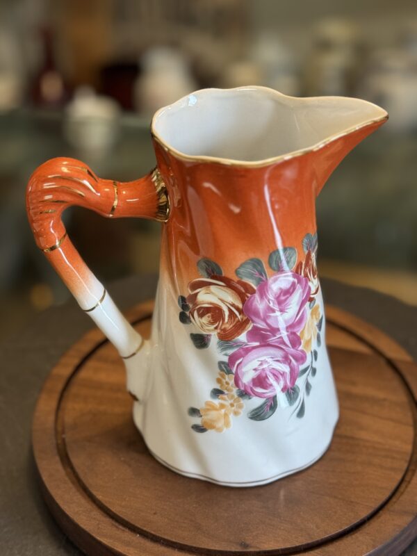 The Vintage Floral Pitcher features a bright orange handle and top, fading to white at the base, with hand-painted pink, yellow, and green flowers on ceramic. Displayed on a round wooden tray against a blurred pottery background.