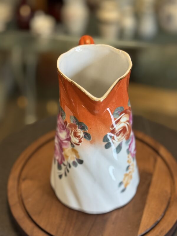 The Vintage Floral Pitcher features a ceramic body with an orange rim and handle, adorned with pink, red, and yellow rose patterns. Displayed on a round wooden tray, it adds a charming touch to any decor.