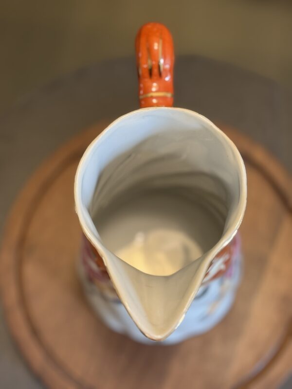 The Vintage Floral Pitcher, featuring a bright orange handle, white interior, wide spout, and decorative floral exterior, rests on a round wooden surface. The overhead view highlights its curved rim and charming details.