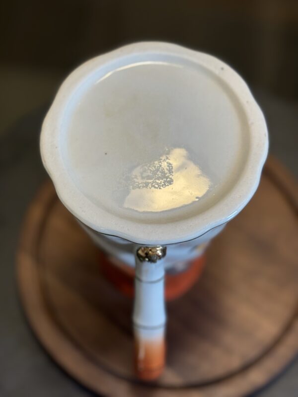 A top-down view of the bottom of a Vintage Floral Pitcher with a curved handle and gold accent, showing its white, glazed ceramic base with shiny, slightly textured areas on a round wooden surface against a softly blurred background.
