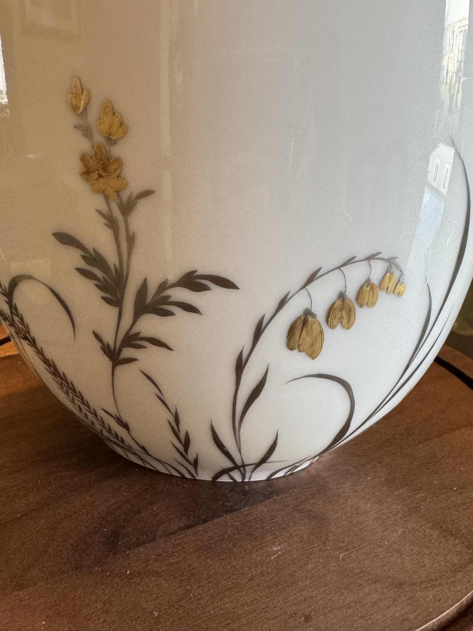 A close-up of a white porcelain vase featuring delicate gold and brown floral designs. The flowers have thin, curved stems and leaves, with some blooms filled in gold. The vase sits on a wooden surface, and soft light reflects off its smooth surface.