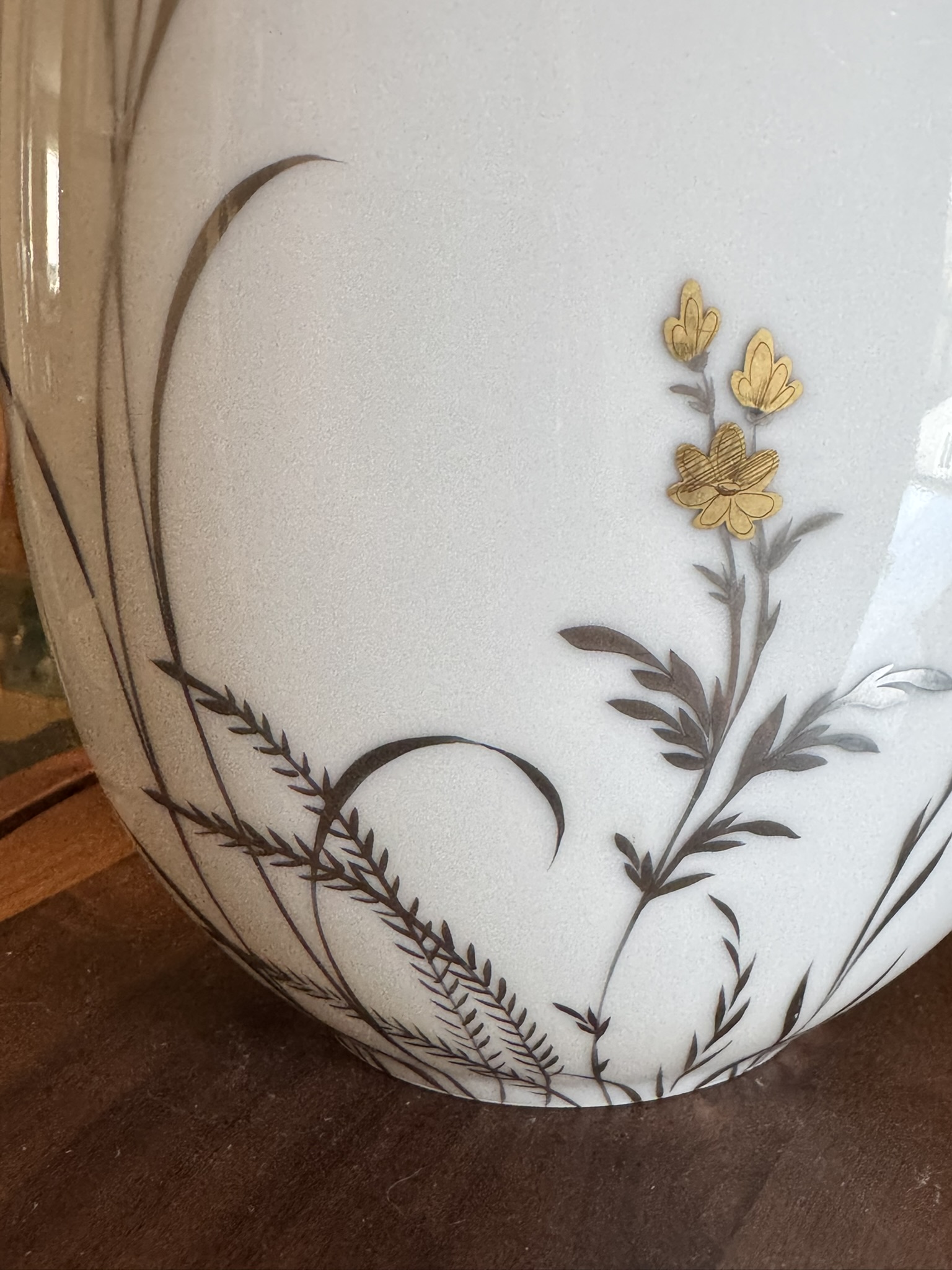 The image shows part of a white vase with delicate black and gold botanical designs. Thin black stems and leaves curve upward, while three gold flowers with intricate detailing stand out near the top right. The vase sits on a wooden surface.