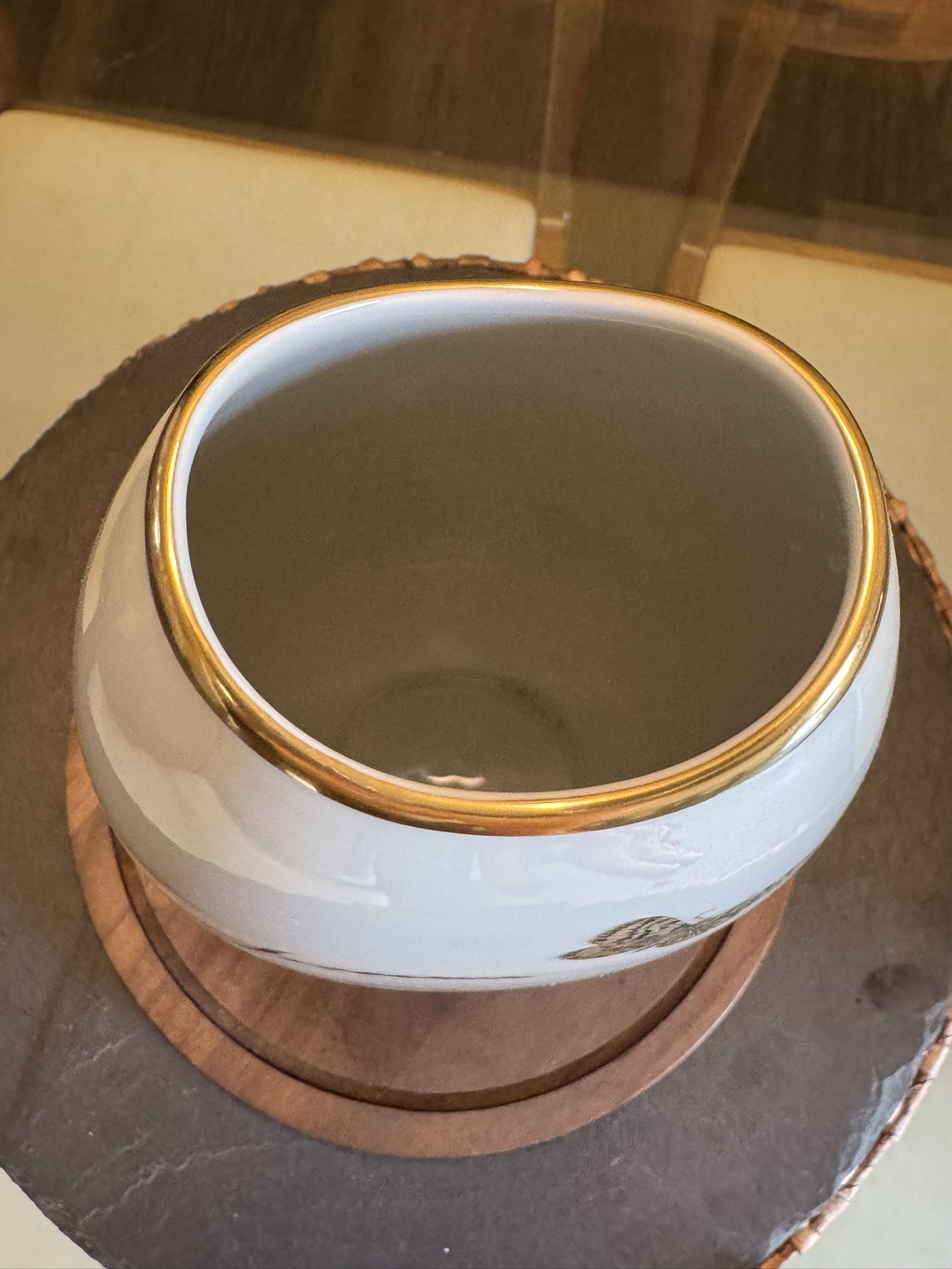 A rounded white ceramic vase with a wide opening and a shiny gold rim sits on a round wooden coaster atop a dark stone surface. The interior appears smooth and clean, and subtle reflections are visible on the glossy edges.