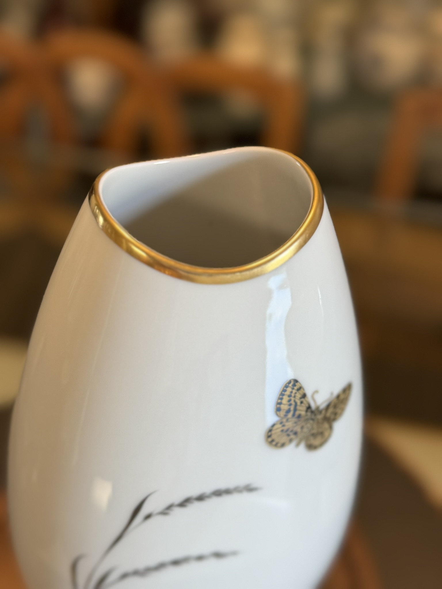A close-up of a glossy white ceramic vase with a wavy, uneven rim outlined in gold. The vase features a small gold butterfly design on its surface. The background is softly blurred, showing brown wooden chairs and a glass table.